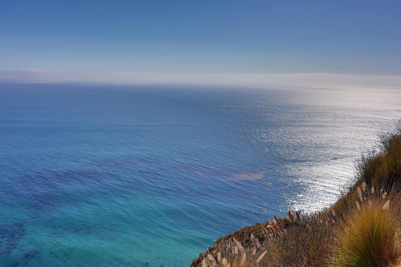 Deep blue ocean seen from a high clifftop, sunlight on the water and grasses in the corner